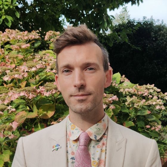 A white man in a cream suit with a floral shirt stands in front of a green bush