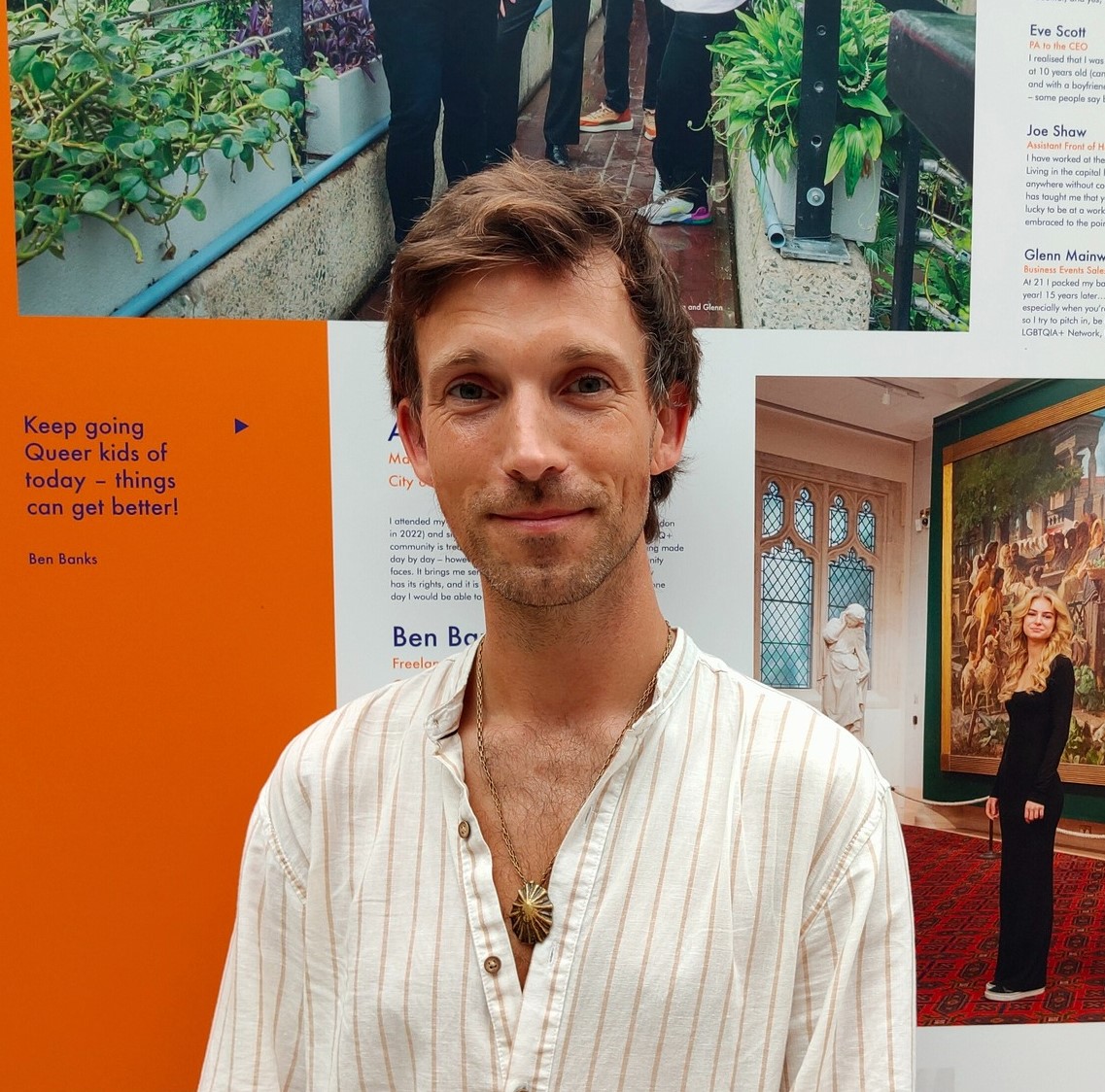 A kind and friendly looking white man in an open topped cream shirt, is shown in front of a queer art exhibition.